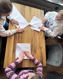  Ein Foto, welches von oben auf einen Tisch herab fotografiert wurde. Zusehen sind 3 Kinder, die auf einem Hoztisch Servietten falten. 