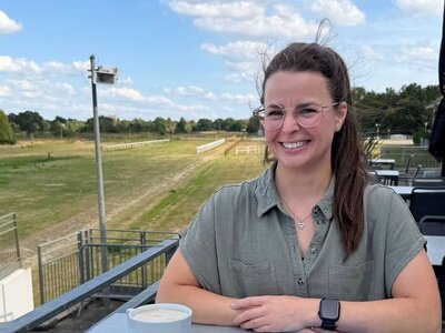 Wencke Bergmann (Project Manager, Sales & Marketing), who has dark hair, is sitting at a table on the terrace. On the table in front of her is a latte, and next to it lies the company newspaper “törn.”