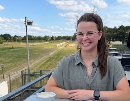 Die dunkelhaarige Wencke Bergmann (Project Manager Sales & Marketing) sitzt an einem Tisch auf der Terrasse. Auf dem Tisch vor ihr steht ein Milchkaffee, daneben liegt die Hauszeitung "törn". 