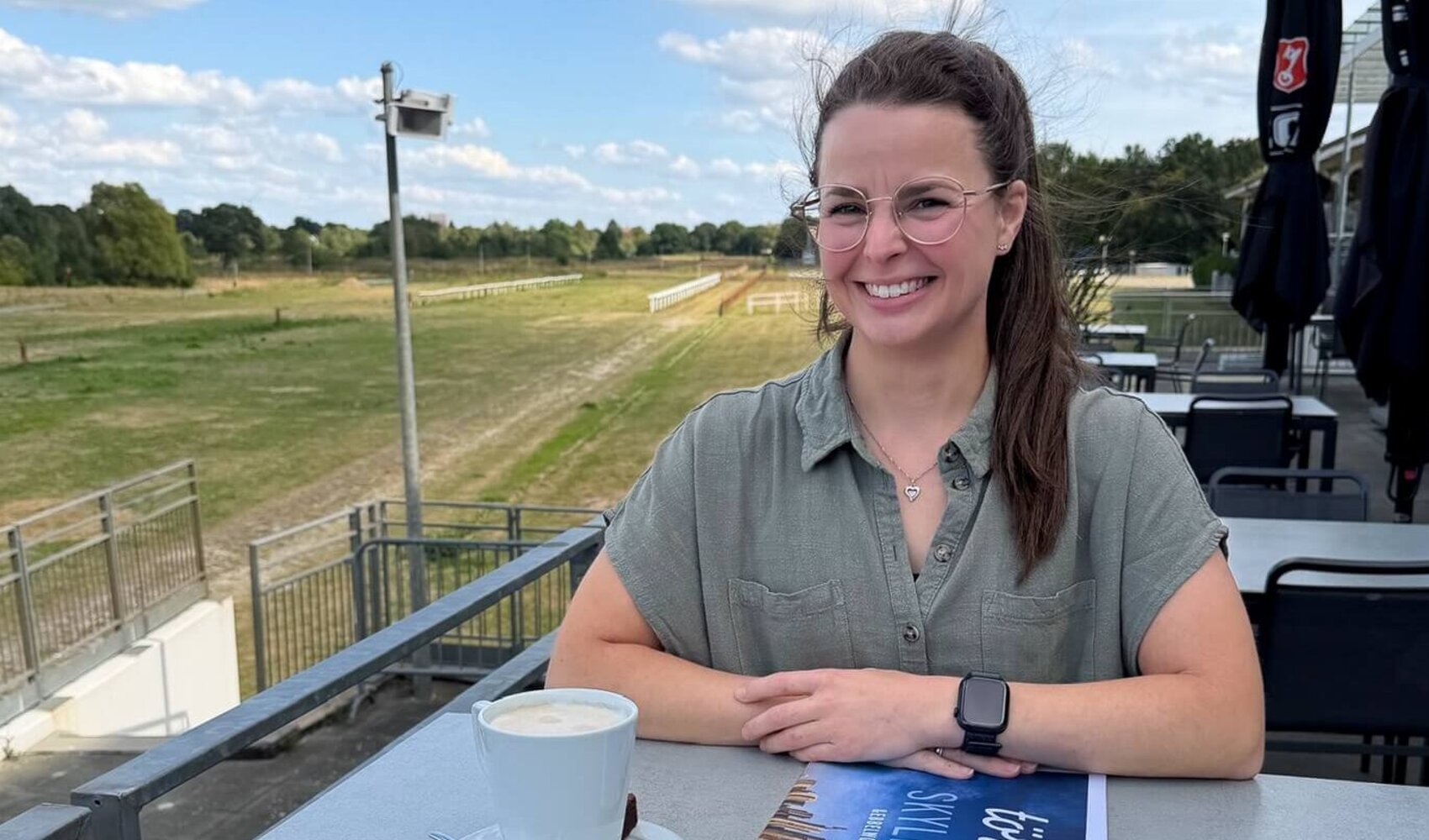 Die dunkelhaarige Wencke Bergmann (Project Manager Sales & Marketing) sitzt an einem Tisch auf der Terrasse. Auf dem Tisch vor ihr steht ein Milchkaffee, daneben liegt die Hauszeitung "törn". 