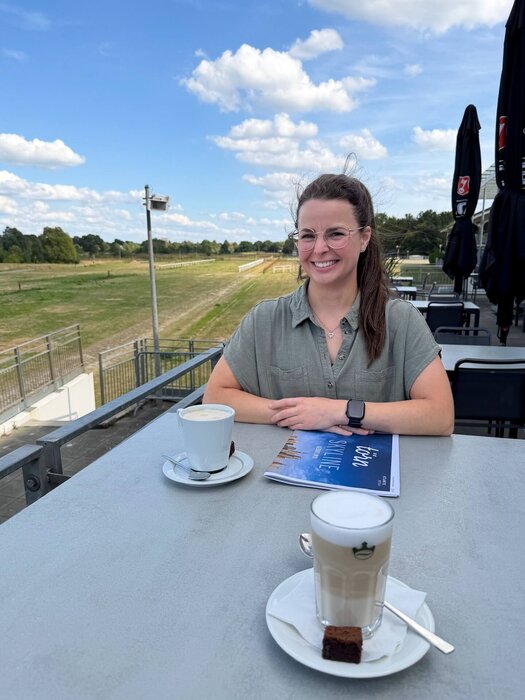 Die dunkelhaarige Wencke Bergmann (Project Manager Sales & Marketing) sitzt an einem Tisch auf der Terrasse. Auf dem Tisch vor ihr steht ein Milchkaffee, daneben liegt die Hauszeitung "törn". 