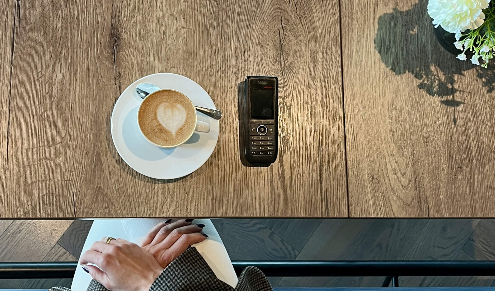 Von oben fotografiert ist die blonde Yasmina Schaller (Rooms Division Managerin) zu sehen, wie sie an einem Tisch auf sitzt. Auf dem Tisch vor ihr steht ein Cappuccino, daneben liegt ein Telefon. 
