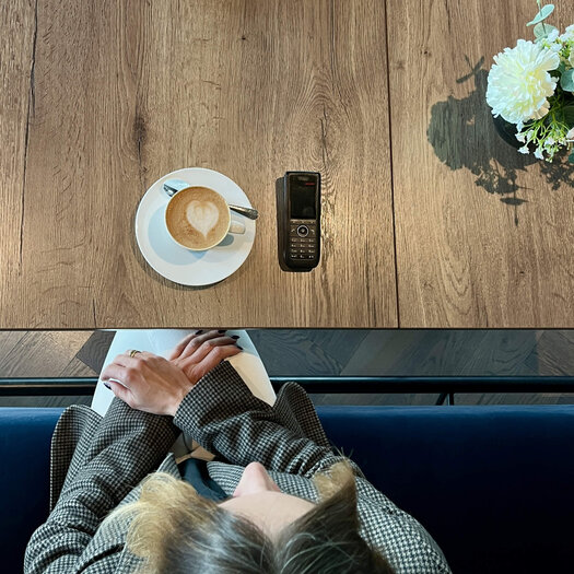 Von oben fotografiert ist die blonde Yasmina Schaller (Rooms Division Managerin) zu sehen, wie sie an einem Tisch auf sitzt. Auf dem Tisch vor ihr steht ein Cappuccino, daneben liegt ein Telefon. 