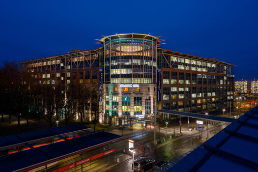 Eine Außenansicht vom ATLANTIC Hotel Airport in Bremen bei Nacht. Links vom Hotel sind viele Bäume zu sehen und vor dem Hotel ist eine Straßenbahnhaltestelle. Im Hotel sind viele beleuchtete Fenster.