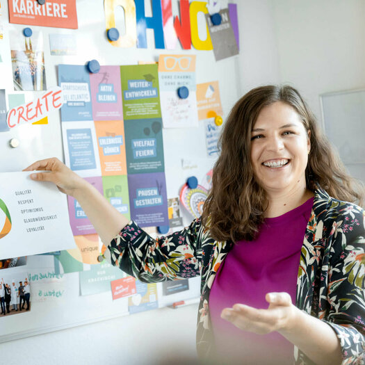 Viktoria Rathjen steht vor ihrem Kreativ-Whiteboard im Büro und hält einen Zettel mit den Unternehmenswerten hoch.