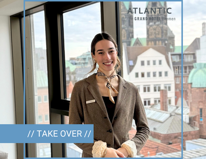 Chiara steht lächelnd vor einem Fenster. Sie trägt einen braunen Blazer, ein beiges Halstuch und ein dunkles Oberteil. Oben rechts in der Ecke ist das Logo der ATANTIC Grand Hotel Bremen abgebildet und unten links ist ein hellblauer Streifen mit der Aufschrift "// TAKE OVER //". Die Person ist umrahmt von hellblauen Linien, die zu einem Quadrat angeordnet sind.