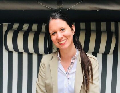 A coffee with Nina Neuhoff (Deputy reservation manager, Severin*s Resort & Spa) Dark-haired Nina Neuhoff sits smiling in a light-colored suit in a striped beach chair. In front of her is a wooden table with a tray holding a latte macchiato, a vase with a pink flower, and a sugar bowl. There is also a potted plant with purple flowers on the table.
