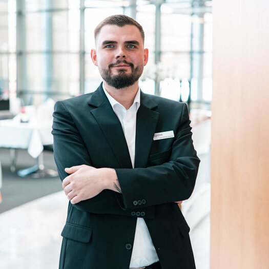 Der dunkelhaarige Jan Niklas Rakoci grinst in die Kamera und hält seine Arme verschränkt. Er trägt ein weißes Hemd, eine schwarze Hose und ein schwarzes Sakko. 