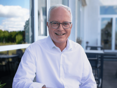 Markus Griesenbeck wears glasses and sits at a table on a terrace wearing a white suit shirt. In front of him is a cappuccino, next to it his smartphone. The blue sky is reflected in the window next to him.