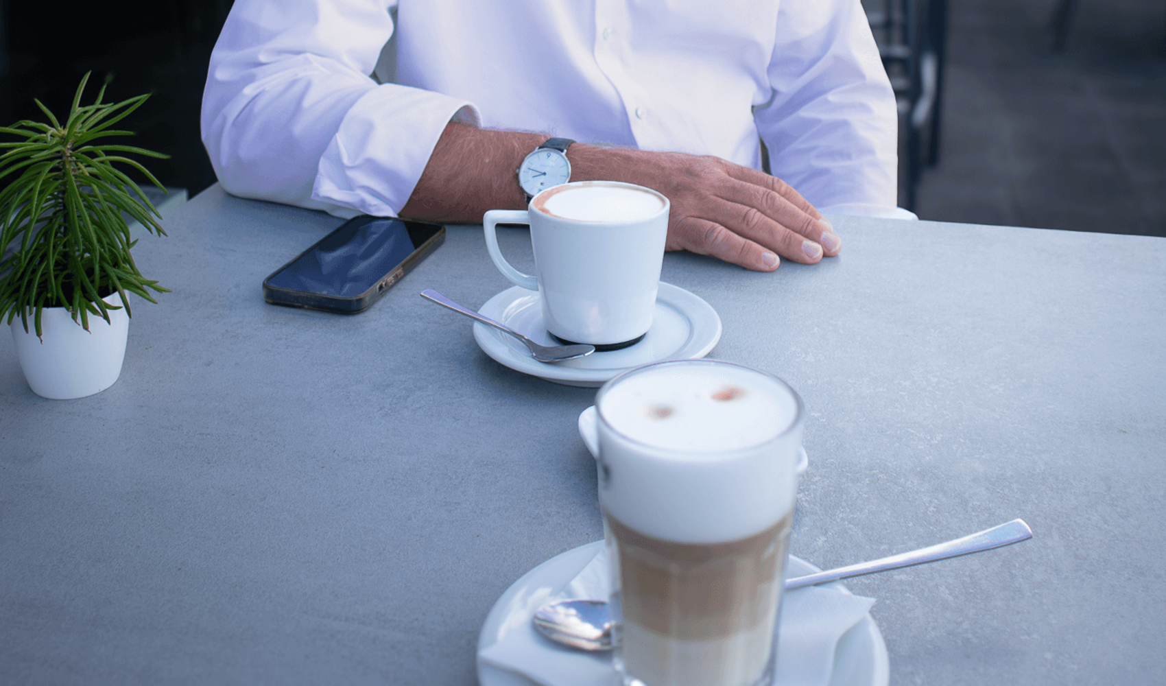 Markus Griesenbeck trägt eine Brille und sitzt in einem weißen Hemd an einem Tisch auf einer Terrasse. Vor ihm steht ein Cappuccino, daneben liegt sein Smartphone. Der blaue Himmel spiegelt sich in der Scheibe neben ihm.
