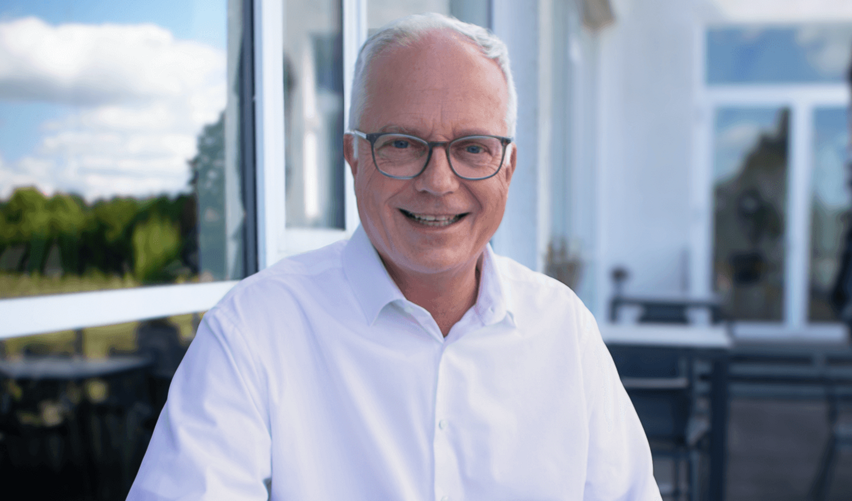 Markus Griesenbeck trägt eine Brille und sitzt in einem weißen Hemd an einem Tisch auf einer Terrasse. Vor ihm steht ein Cappuccino, daneben liegt sein Smartphone. Der blaue Himmel spiegelt sich in der Scheibe neben ihm.