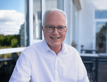 A coffee with Markus Griesenbeck (CEO, ATLANTIC Hotels) Markus Griesenbeck wears glasses and sits at a table on a terrace wearing a white suit shirt. In front of him is a cappuccino, next to it his smartphone. The blue sky is reflected in the window next to him.