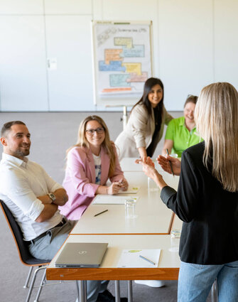 Eine Trainerin mit blonden Haaren steht in einem Seminarraum vor einem Blocktisch an dem neun Teilnehmende sitzen und interessiert in ihre Richtung schauen.