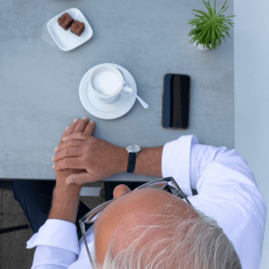 Von oben fotografiert ist Markus Griesenbeck in einem weißen Hemd zu sehen, wie er an einem grauen Tisch sitzt. Seine Hände liegen übereinander auf dem Tisch. Vor ihm steht ein Cappuccino, ein kleiner Teller mit Brownies und eine grüne Pflanze. Sein Smartphone liegt ebenfalls auf dem Tisch.