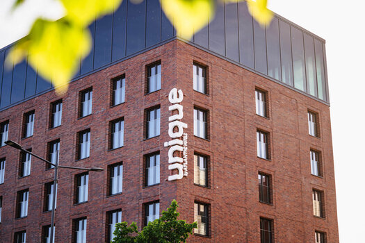 Eine Außenansicht des unique by ATLANTIC Hotels Kiel. Das Gebäude hat viele Fenster und in der obersten Etage ist eine große Fensterfront. Auf dem Gebäude steht "unique by ATLANTIC Hotels".