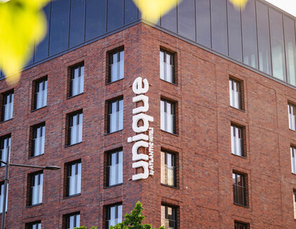 An exterior view of the unique by ATLANTIC Hotel Kiel. The building has many windows and there is a large window front on the top floor. The building is labeled “unique by ATLANTIC Hotels”.