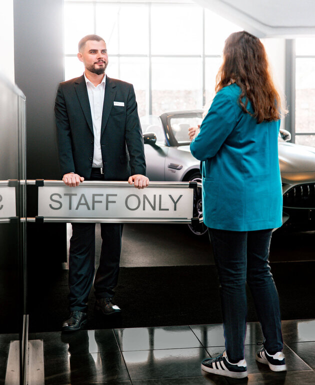 Der dunkelhaarige Jan Niklas Rakoci steh hinter einem Schild auf dem "Staff only" steht. Vor ihm steht eine Frau mit dunklen Haaren und einem türkisen Blazer. Im Hintergrund steht ein silbernes Auto. Er trägt ein weißes Hemd, eine schwarze Hose und ein schwarzes Sakko. 