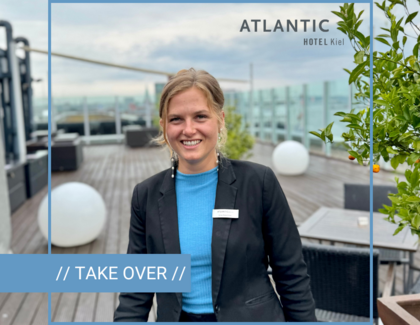Eine Frau steht lächelnd auf einer große Terrasse. Sie trägt eine schwarze Hose, einen schwarzen Blazer und eine blaues Oberteil. Oben rechts in der Ecke ist das Logo des ATANTIC Hotel Kiel abgebildet und unten links ist ein hellblauer Streifen mit der Aufschrift "// TAKE OVER //". Die Person ist umrahmt von hellblauen Linien, die zu einem Quadrat angeordnet sind. 