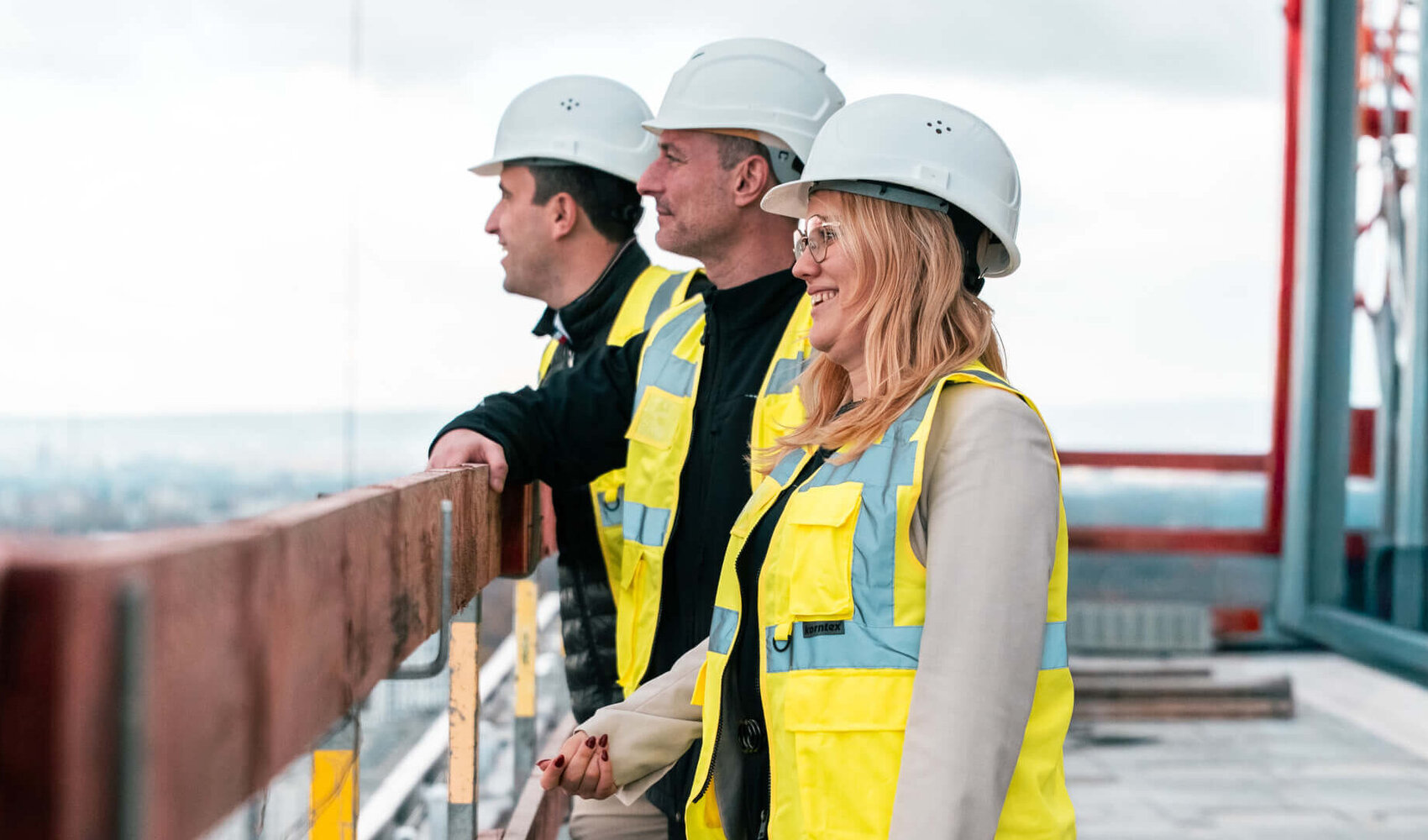 Teambild ATLANTIC Hotel Frankfurt auf der Baustelle Zwei Männer und eine Frau mit Bauhelmen und Sicherheitswesten schauen über die Dachbrüstung in die Ferne.