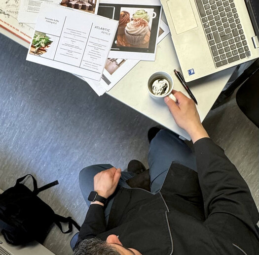Auf einen Kaffee mit Patrick Beainy (Küchenchef, ATLANTIC Hotel Frankfurt) Von oben fotografiert ist der dunkelhaarige Patrick Beainy zu sehen, wie er an einem Schreibtisch sitzt. In den Händen hält er einen Kaffee und auf dem Tisch liegen zudem ein Laptop und einige Zettel.