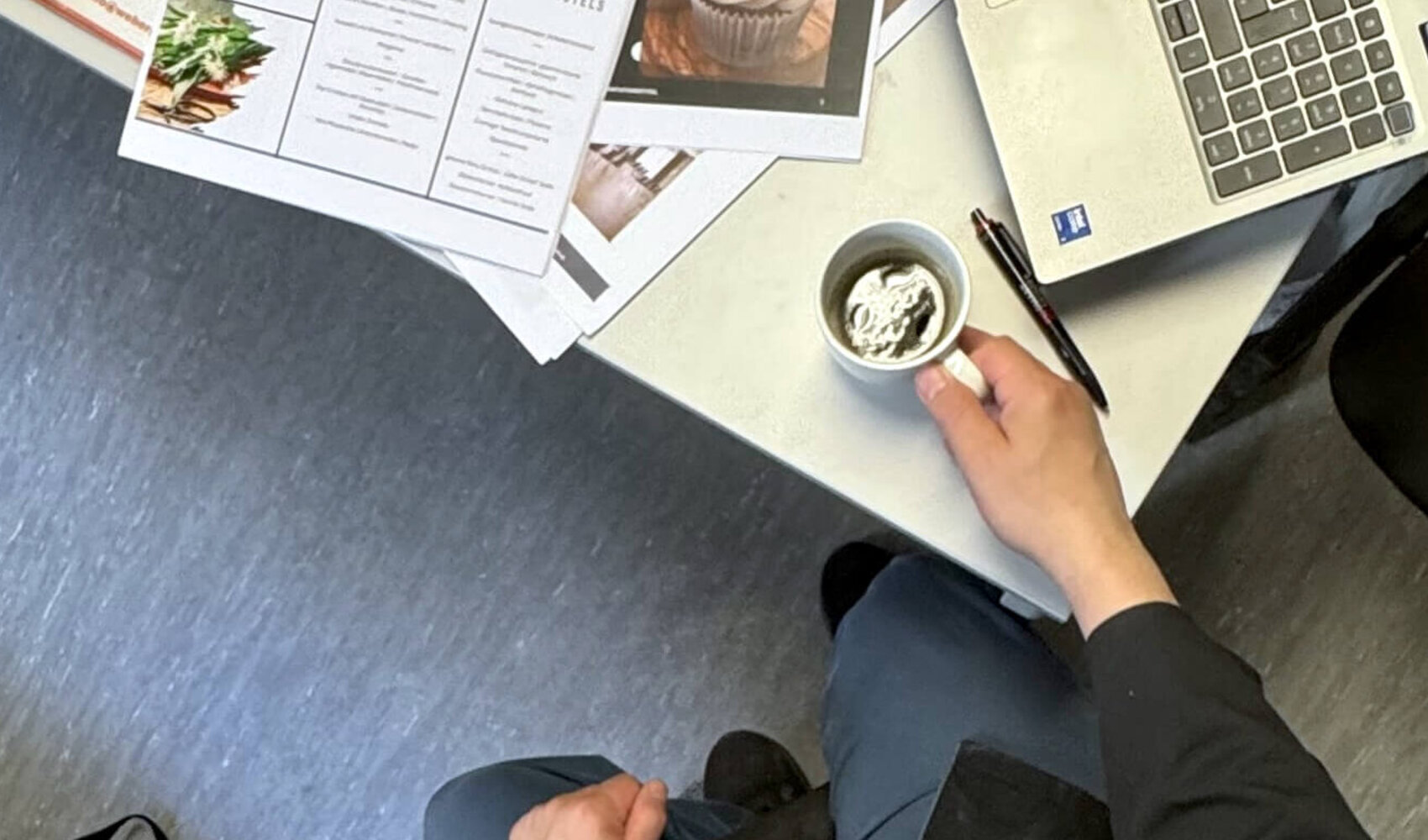 Auf einen Kaffee mit Patrick Beainy (Küchenchef, ATLANTIC Hotel Frankfurt) Von oben fotografiert ist der dunkelhaarige Patrick Beainy zu sehen, wie er an einem Schreibtisch sitzt. In den Händen hält er einen Kaffee und auf dem Tisch liegen zudem ein Laptop und einige Zettel.