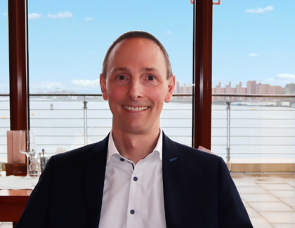 A coffee with hotel director Florian Schönwetter Hotel manager Florian Schönwetter wears a suit and sits smiling at a wooden restaurant table with a light-colored table runner. His arms are resting on the backs of the chairs. On the table is a cappuccino and a latte macchiato - each with a brownie on the saucer - and a small vase with a flower. A mobile phone rests on a blue notebook. The Wilhelmshaven harbor basin can be seen in the background under a blue sky.