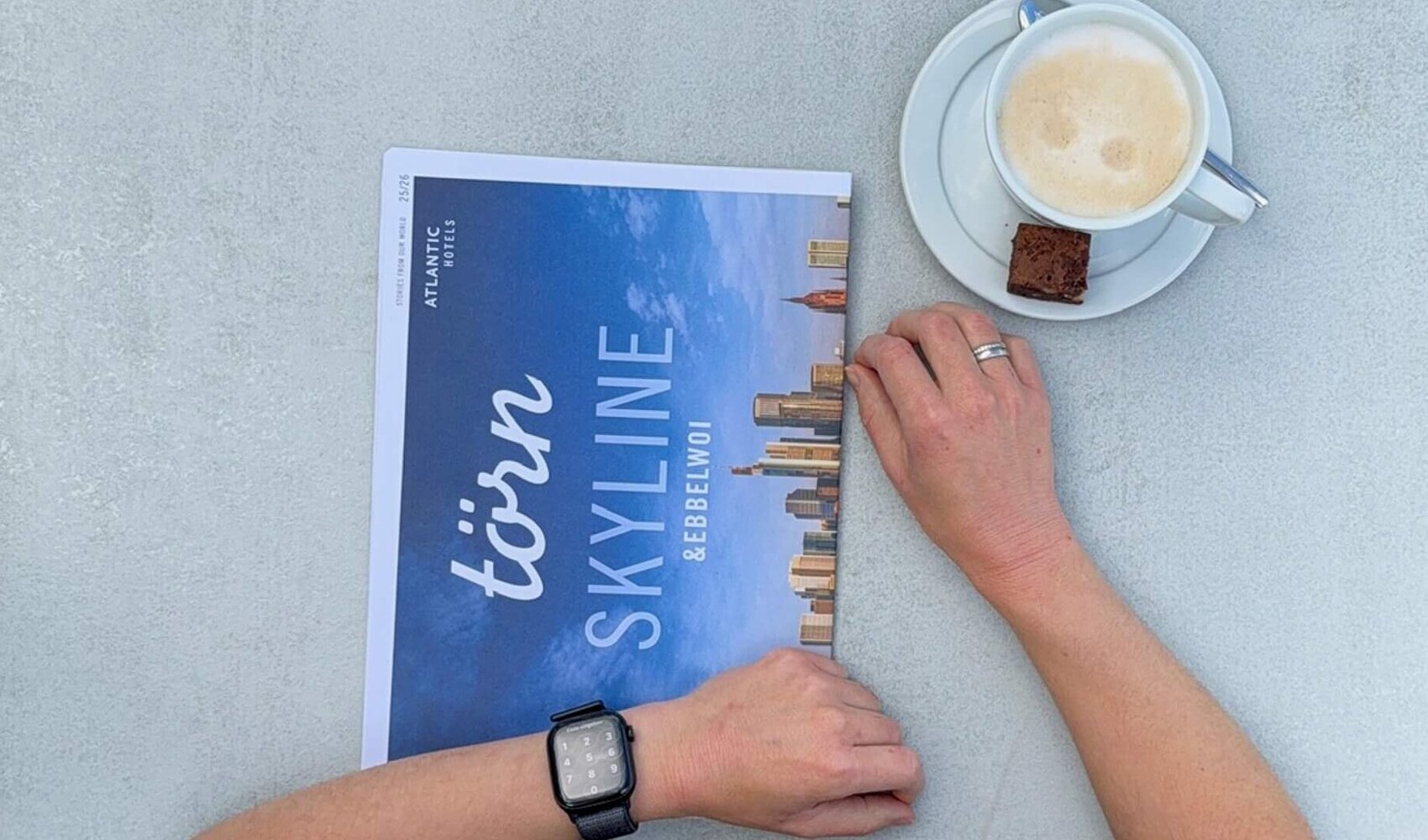 Von oben fotografiert ist die dunkelhaarige Wencke Bergmann (Project Manager Sales & Marketing) zu sehen, wie sie an einem Tisch auf der Terrasse sitzt. Auf dem Tisch vor ihr steht ein Milchkaffee, daneben liegt die Hauszeitung "törn". 