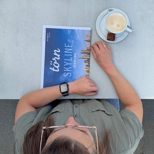 Von oben fotografiert ist die dunkelhaarige Wencke Bergmann (Project Manager Sales & Marketing) zu sehen, wie sie an einem Tisch auf der Terrasse sitzt. Auf dem Tisch vor ihr steht ein Milchkaffee, daneben liegt die Hauszeitung "törn". 