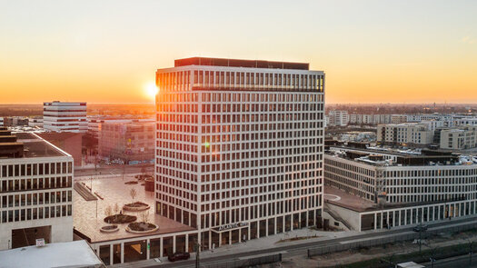 An exterior view of the ATLANTIC Hotel Heidelberg. The hotel is beige, very tall and has many windows. "ATLANTIC Hotels" is written above the hotel entrance. A city and a sunset can be seen in the background.