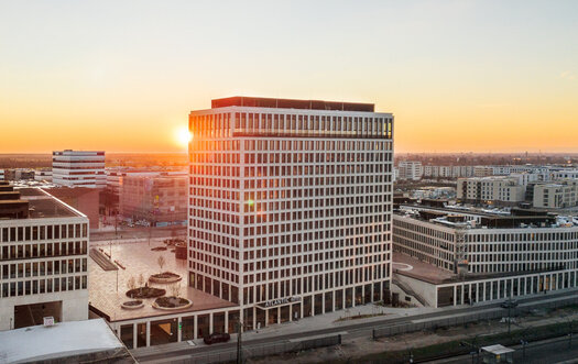 An exterior view of the ATLANTIC Hotel Heidelberg. The hotel is beige, very tall and has many windows. "ATLANTIC Hotels" is written above the hotel entrance. A city and a sunset can be seen in the background.