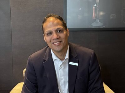 Der dunkelhaarige Amr Ahmed sitzt auf einem beigen Stuhl. Er trägt einen schwarzen Anzug, ein weißes Hemd. Vor ihm steht ein Tisch auf welchem zwei Kaffee, eine Blume und ein Teller mit kleinen Brownies steht. Die Wand hinter ihm ist schwarz. 