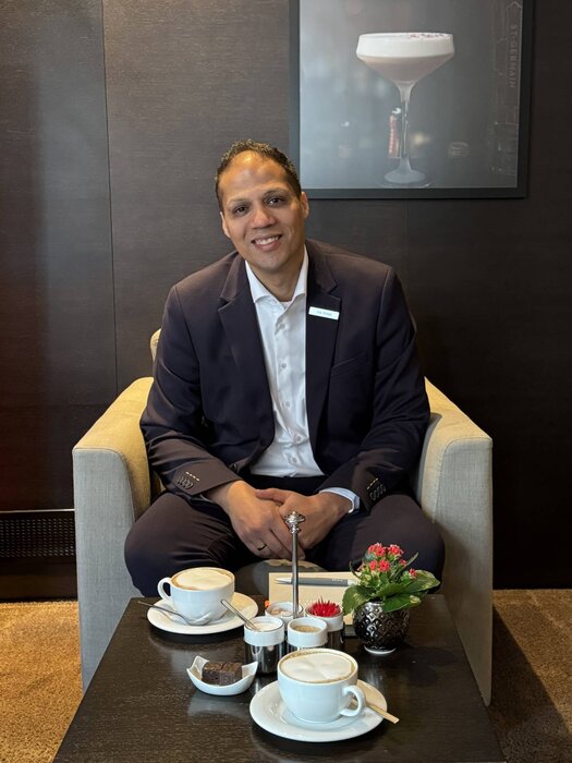 Auf einen Kaffee mit Front Office Manager Amr Ahmed Der dunkelhaarige Amr Ahmed sitzt auf einem beigen Stuhl. Er trägt einen schwarzen Anzug, ein weißes Hemd. Vor ihm steht ein Tisch auf welchem zwei Kaffee, eine Blume und ein Teller mit kleinen Brownies steht. Die Wand hinter ihm ist schwarz.