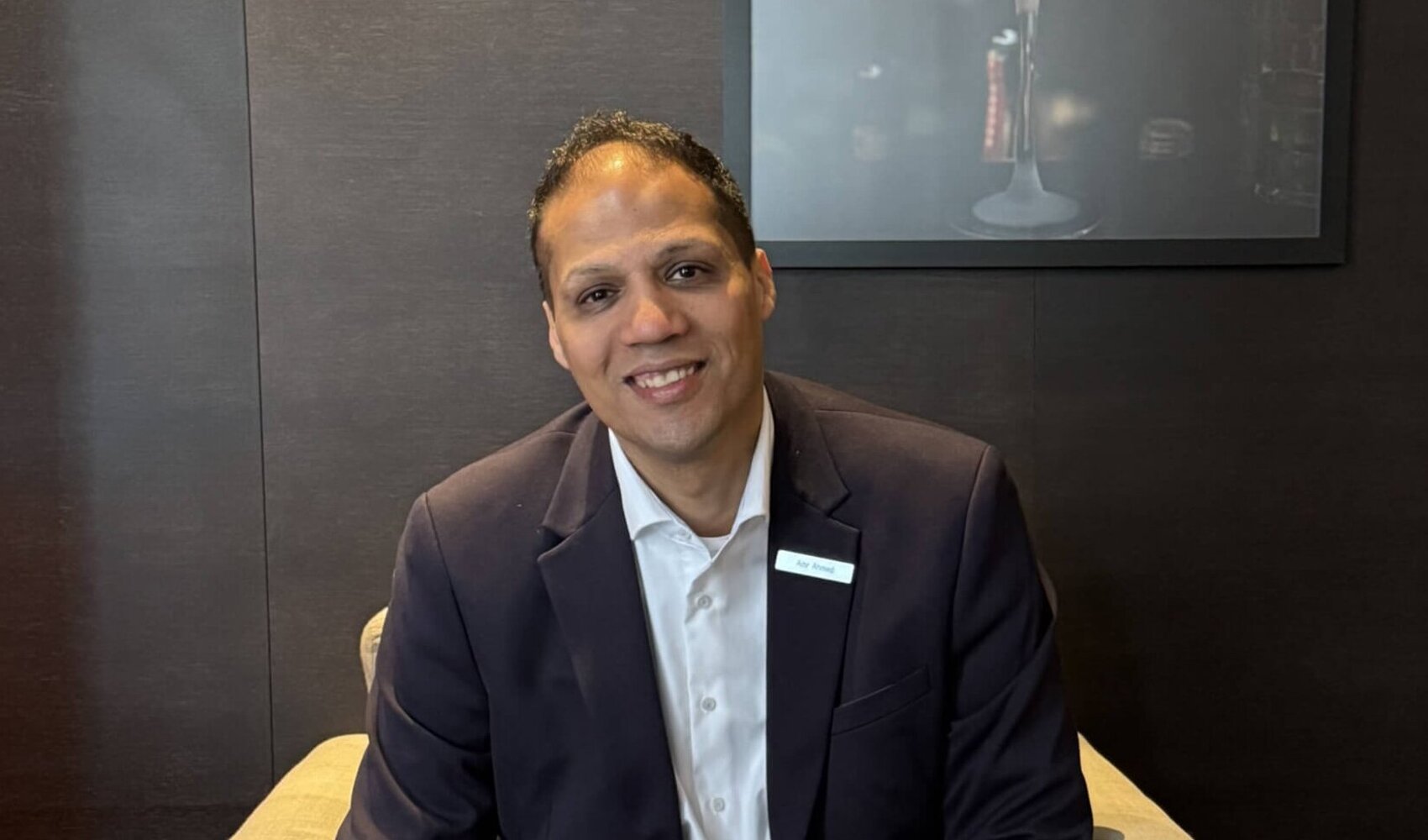 Auf einen Kaffee mit Front Office Manager Amr Ahmed Der dunkelhaarige Amr Ahmed sitzt auf einem beigen Stuhl. Er trägt einen schwarzen Anzug, ein weißes Hemd. Vor ihm steht ein Tisch auf welchem zwei Kaffee, eine Blume und ein Teller mit kleinen Brownies steht. Die Wand hinter ihm ist schwarz.