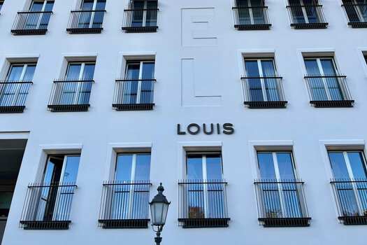 Eine Außenansicht des LOUIS Hotel in München. Das Hotel hat eine weiße Außenfassade, viele Fenster und die Aufschrift "LOUIS". 