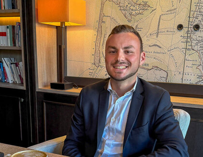 Auf einen Kaffee mit Cem Eryaman (F&B Assistant, Severin*s Resort & Spa) Der dunkelhaarige Cem Eryamansitzt glücklich an einem Holztisch. Auf dem Tisch vor ihm stehen zwei Cappuccini sowie zwei Wassergläser. Ein mobiles Telefon liegt ebenfalls auf dem Tisch. Im Hintergrund sind ein Holzschrank und eine Weltkarte zu sehen.