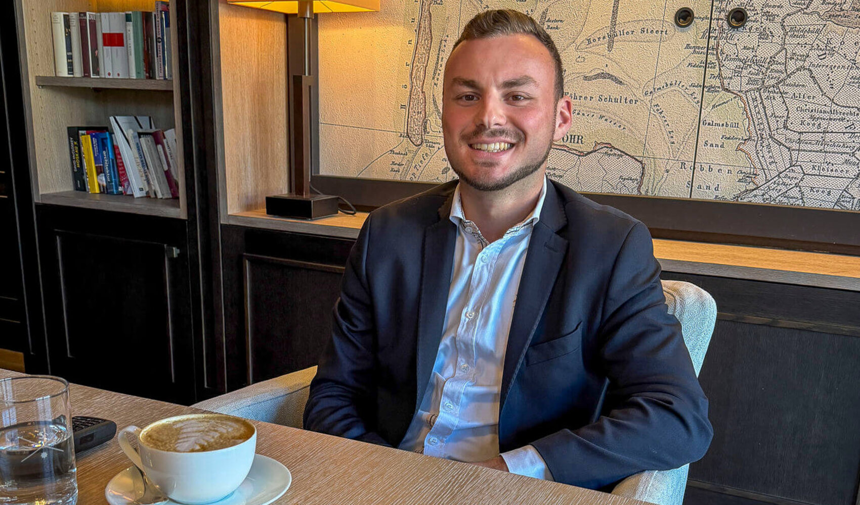 Auf einen Kaffee mit Cem Eryaman (F&B Assistant, Severin*s Resort & Spa) Der dunkelhaarige Cem Eryamansitzt glücklich an einem Holztisch. Auf dem Tisch vor ihm stehen zwei Cappuccini sowie zwei Wassergläser. Ein mobiles Telefon liegt ebenfalls auf dem Tisch. Im Hintergrund sind ein Holzschrank und eine Weltkarte zu sehen.