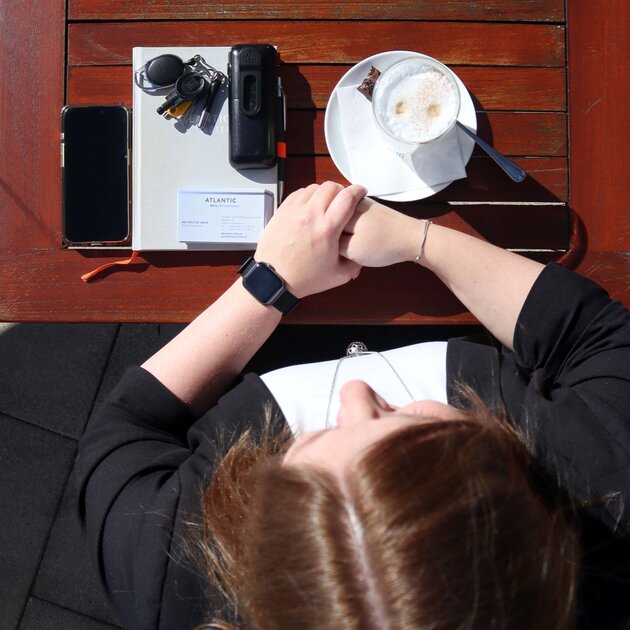 Von oben fotografiert ist die dunkelhaarige Ann-Christine zu sehen wie sie an einem braunen Holztisch sitzt. Ihre Hände liegen übereinander auf dem Tisch. Ein Latte Macchiato, ein Telefon, ein Buch, Schlüssel und ein Handy liegen ebenfalls auf dem Tisch. 