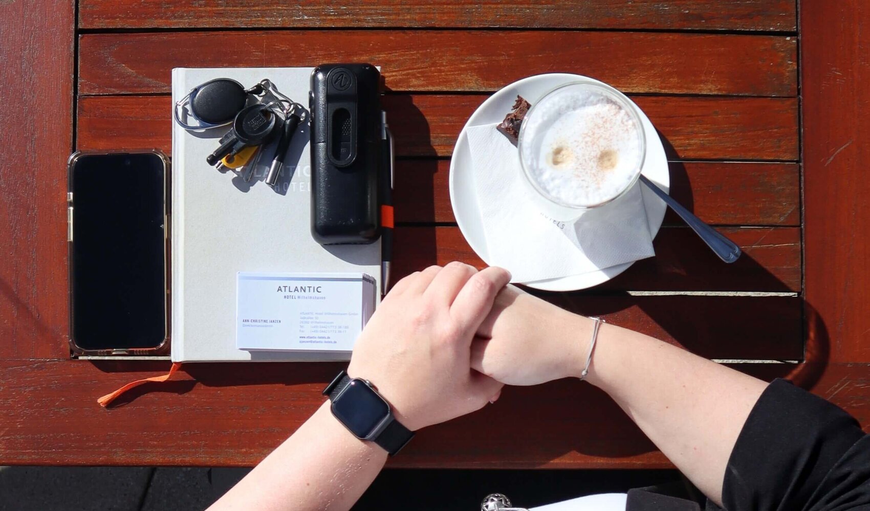 Von oben fotografiert ist die dunkelhaarige Ann-Christine zu sehen wie sie an einem braunen Holztisch sitzt. Ihre Hände liegen übereinander auf dem Tisch. Ein Latte Macchiato, ein Telefon, ein Buch, Schlüssel und ein Handy liegen ebenfalls auf dem Tisch. 