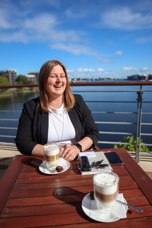 Die dunkelhaarige Ann-Christine Janzen sitzt glücklich lachend auf der Hotelterrasse an einem braunen Holztisch. Auf dem Tisch vor ihr stehen zwei Latte Macchiato. Zudem liegt ein Notizbuch, ein mobiles Telefon, ein Handy, Visitenkarten und ein Schlüssel auf dem Tisch.