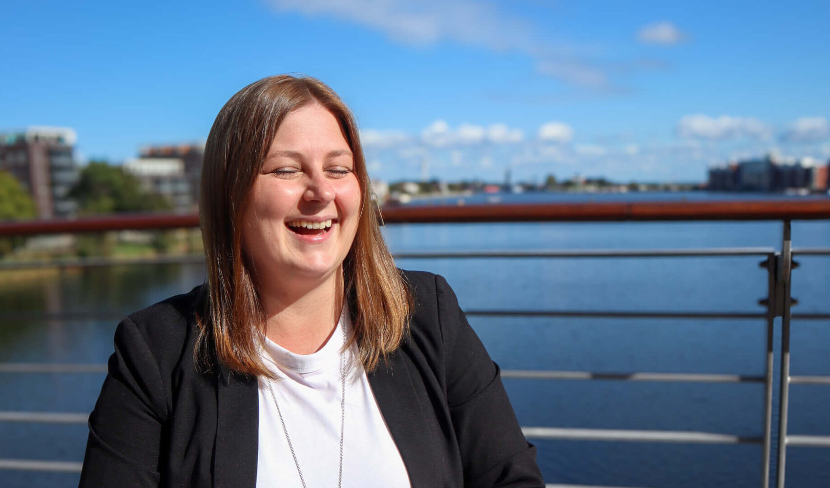 Auf einen Kaffee mit Ann-Christine Janzen (Direktionsassistentin, ATLANTIC Hotel Wilhelmshaven) Die dunkelhaarige Ann-Christine Janzen sitzt glücklich lachend auf der Hotelterrasse an einem braunen Holztisch. Auf dem Tisch vor ihr stehen zwei Latte Macchiato. Zudem liegt ein Notizbuch, ein mobiles Telefon, ein Handy, Visitenkarten und ein Schlüssel auf dem Tisch.