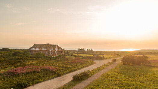 Außenansicht Landhaus Severin*s Morsum Kliff, Sylt Eine Außenansicht des Landhaus Severin*s Morsum Kliff auf Sylt. Ein Gebäude ist umgeben von viel Natur und in der Nähe des Meeres. Zwei große Flaggen wehen vor dem Hotel und das Gebäude hat ein Reetdach.