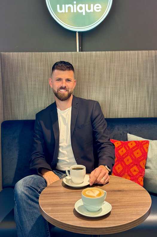 Frederik Schönke sitzt auf einer Bank an einem Tisch. Vor ihm steht ein schwarzer Kaffee und ein Cappuccino auf einem Tisch.