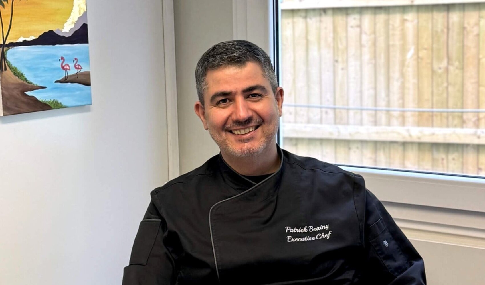 Auf einen Kaffee mit Patrick Beainy (Küchenchef, ATLANTIC Hotel Frankfurt) Der dunkelhaarige Patrick Beainy sitzt an seinem Schreibtisch in einem Baucontainer und lacht in die Kamera. Vor ihm steht eine Tasse Kaffee. Im Hintergrund hängt ein buntes Bild mit Wasser und Palmen an der Wand. Er trägt eine schwarze Küchenuniform.