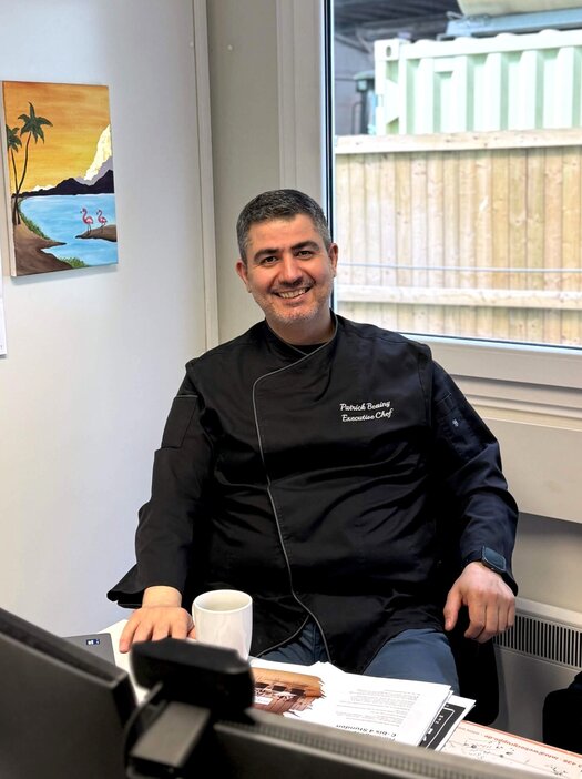 Auf einen Kaffee mit Patrick Beainy (Küchenchef, ATLANTIC Hotel Frankfurt) Der dunkelhaarige Patrick Beainy sitzt an seinem Schreibtisch in einem Baucontainer und lacht in die Kamera. Vor ihm steht eine Tasse Kaffee. Im Hintergrund hängt ein buntes Bild mit Wasser und Palmen an der Wand. Er trägt eine schwarze Küchenuniform.