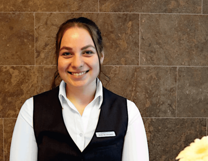 Die dunkelhaarige Empfangsmitarbeiterin Deike Borchmann lächelt in die Kamera während sie in ihrer Rezeptionsuniform an einem Tisch mit weißer Tischdecke sitzt. Auf dem Tisch stehen zwei Latte Macchiato, ein Zuckerpott und eine Blume in einer kleinen Vase. Im Hintergrund ist eine geflieste Wand zu sehen. 