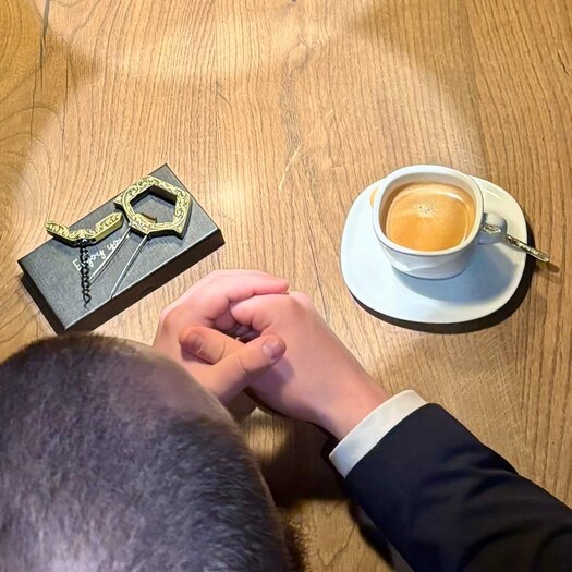 Von oben fotografiert ist Leon Decker sitzend an einem Holztisch zu sehen. Auf dem Tisch steht ein Kaffee Crema, daneben liegen seine gefalteten Hände und ein Durand-Korkenzieher.
