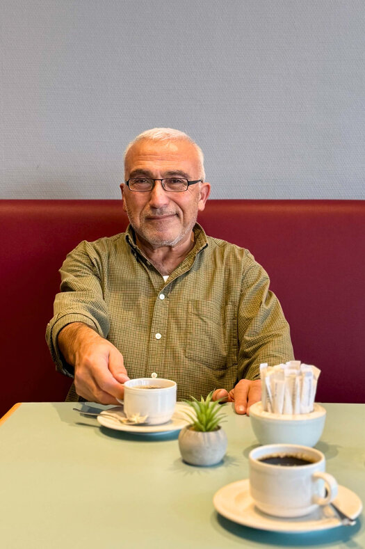 Auf einen Kaffee mit Ali Güney (Housekeeping Allrounder, ATLANTIC Hotel Vegesack) Ali Güney trägt eine Brille und ein grün gemustertes Hemd, während er auf einer rot gepolsterten Sitzbank sitzt. In seiner rechten Hand hält er eine Kaffeetasse, die vor ihm auf dem Tisch steht. Auf dem Tisch steht noch eine weitere Tasse mit Kaffee, eine kleine Pflanze und ein Behälter mit abgepacktem Zucker