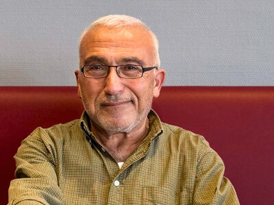 Ali Güney trägt eine Brille und ein grün gemustertes Hemd, während er auf einer rot gepolsterten Sitzbank sitzt. In seiner rechten Hand hält er eine Kaffeetasse, die vor ihm auf dem Tisch steht. Auf dem Tisch steht noch eine weitere Tasse mit Kaffee, eine kleine Pflanze und ein Behälter mit abgepacktem Zucker