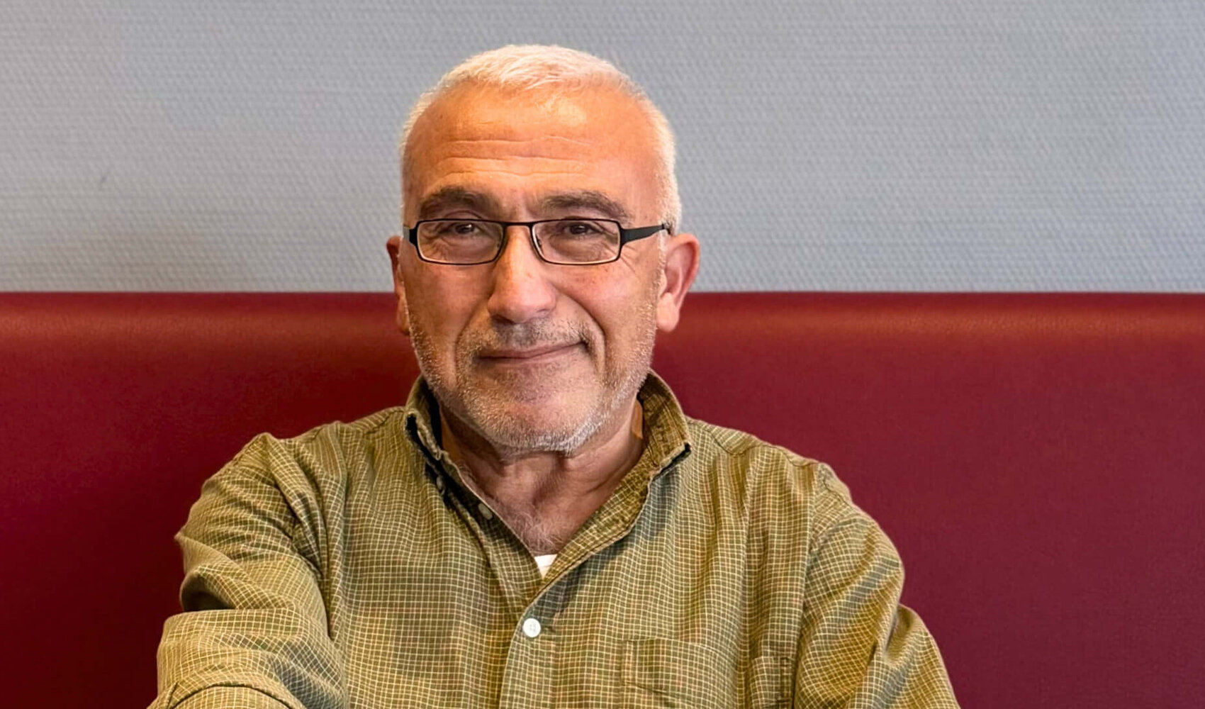 Auf einen Kaffee mit Ali Güney (Housekeeping Allrounder, ATLANTIC Hotel Vegesack) Ali Güney trägt eine Brille und ein grün gemustertes Hemd, während er auf einer rot gepolsterten Sitzbank sitzt. In seiner rechten Hand hält er eine Kaffeetasse, die vor ihm auf dem Tisch steht. Auf dem Tisch steht noch eine weitere Tasse mit Kaffee, eine kleine Pflanze und ein Behälter mit abgepacktem Zucker