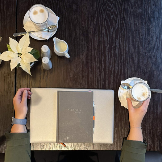 A coffee with mit Laura Sophie Keller Photographed from above, Laura's hands can be seen as she sits at a brown wooden table. Her hands are resting on the table to the right and left of her laptop. Two coffees, a flower, a notebook, and milk are also on the table.